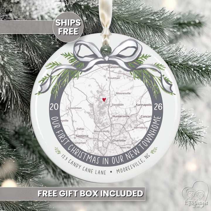 A round white Christmas ornament with a printed map, green pine branches, and a gray ribbon bow design, hanging against snowy tree branches. Text overlays read &