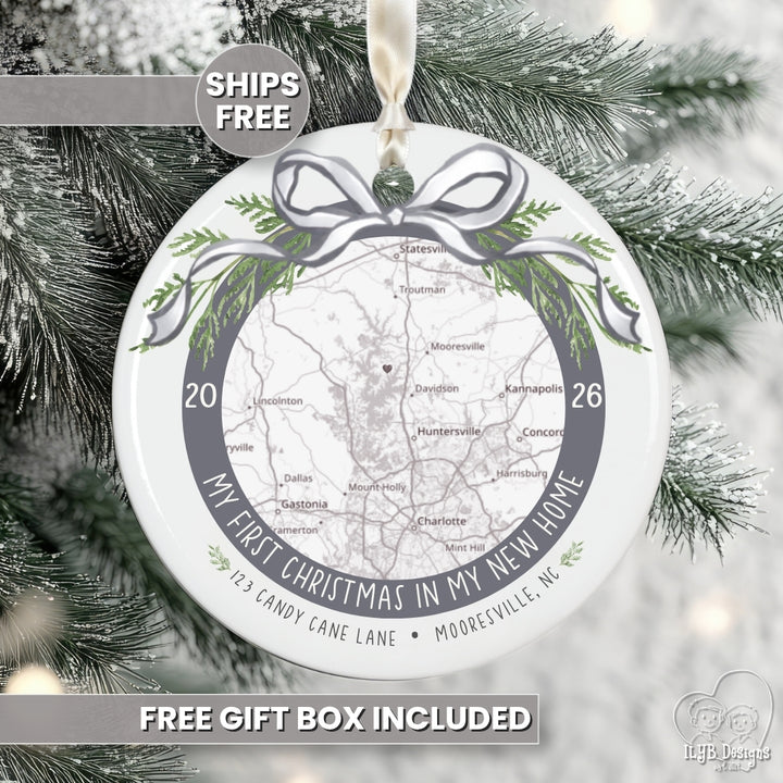 A round white new home Christmas ornament with a printed map, green pine branches, and a gray ribbon bow design, hanging against snowy tree branches. Text overlays read &
