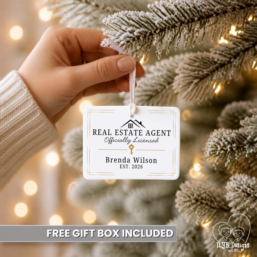 A hand wearing a cream knit sleeve hangs a white rectangular ornament tag from a flocked Christmas tree branch with warm string lights in the background. The tag reads &
