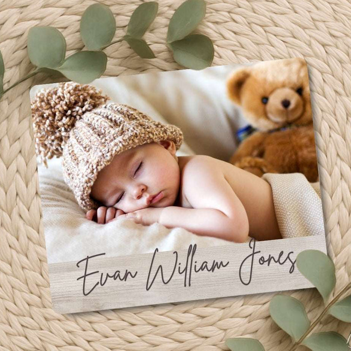 Newborn baby in a knitted hat with a teddy bear, lying on a wooden board with engraved name, surrounded by green leaves.