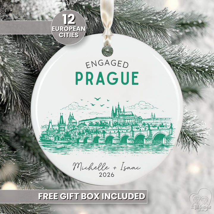 A round white Christmas ornament with a green line-art illustration of the Prague skyline, hanging from a white ribbon against snowy pine branches. The ornament reads &