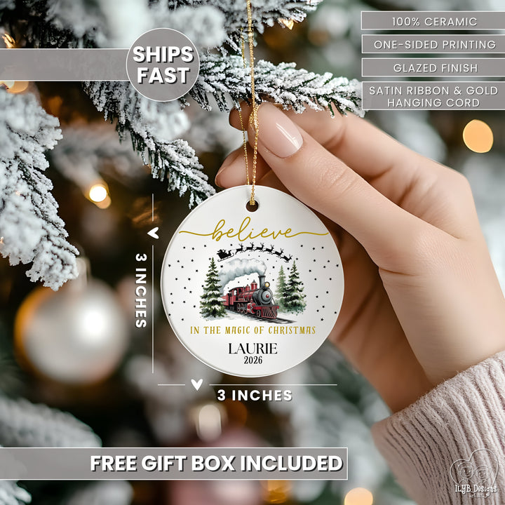 Hand hanging "believe in the magic of Christmas" red express train ornament on a Christmas tree and showing dimensions of the ornament in perspective next to the hand. Ornament dimensions are 3 inches x 3 inches. Other features are highlighted: 100% ceramic, sublimation printing, one-sided print, glazed finish, gold hanging cord, free gift box included, ships fast.