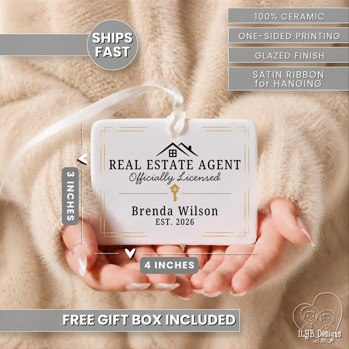A personalized ceramic real estate agent ornament held in two hands against a soft beige faux fur background. The ornament is white with black text and a small gold key graphic, with a white satin ribbon hanging from the top.