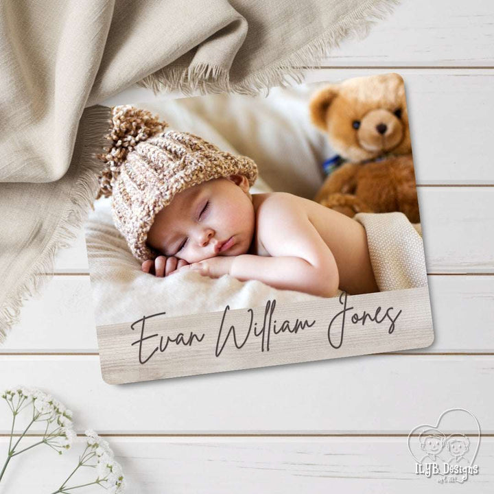 Newborn baby sleeping on a wooden board with 'Evan William Jones' engraved, surrounded by a teddy bear and blanket.