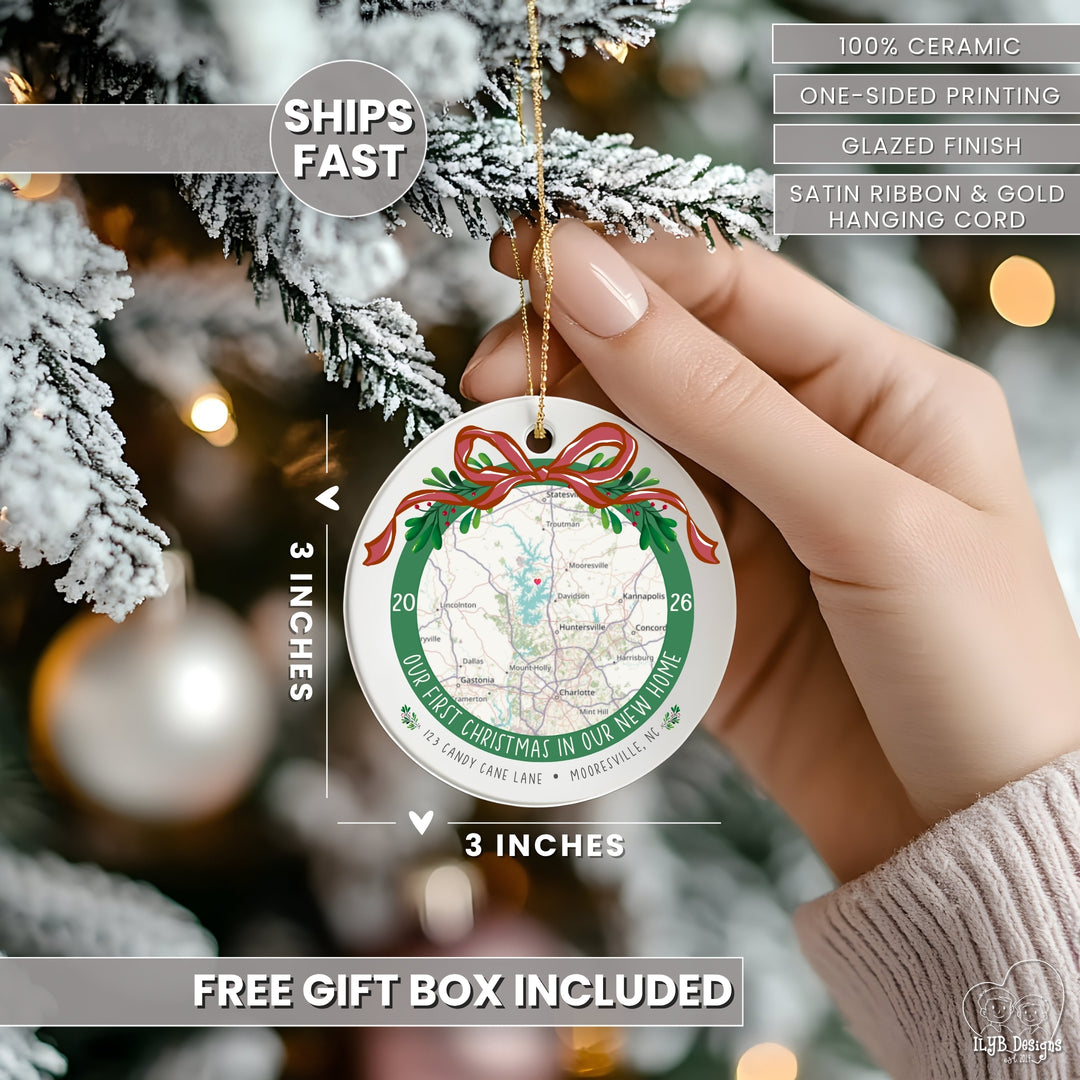 A hand holds a round ceramic Christmas ornament against a blurred Christmas tree background. The ornament is white with a printed map design, green border text, red bow and holly accents, and a gold hanging cord.