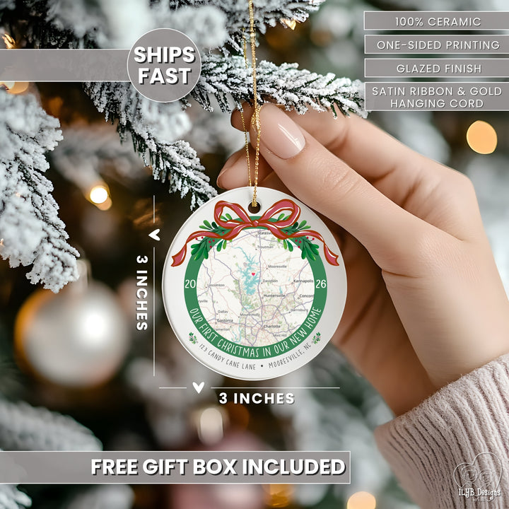 A hand holds a round ceramic Christmas ornament against a blurred Christmas tree background. The ornament is white with a printed map design, green border text, red bow and holly accents, and a gold hanging cord.