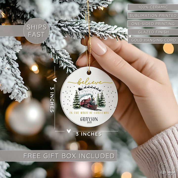 Hand hanging "believe in the magic of Christmas" red express train ornament on a Christmas tree and showing dimensions of the ornament in perspective next to the hand. Ornament dimensions are 3 inches x 3 inches. Other features are highlighted: 100% ceramic, sublimation printing, one-sided print, glazed finish, gold hanging cord, free gift box included, ships fast.