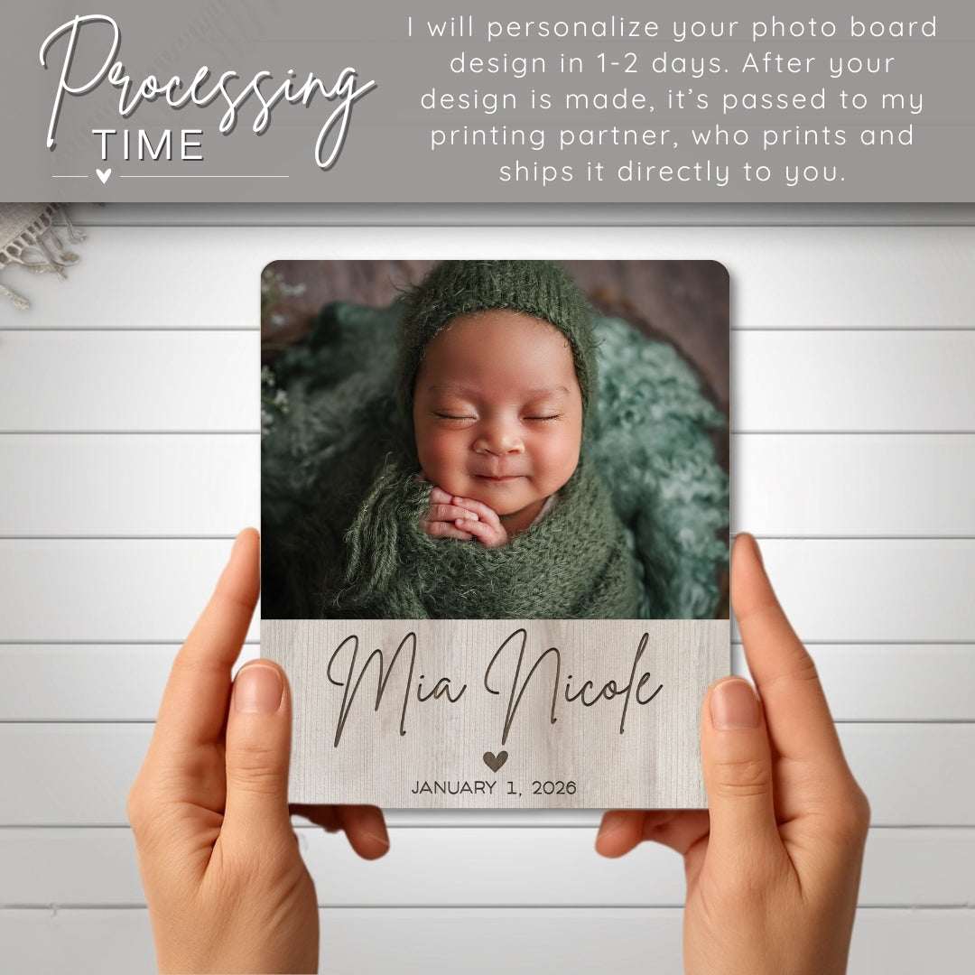 Person holding a photo board with a baby's name and date printed on it, against a light wooden background.