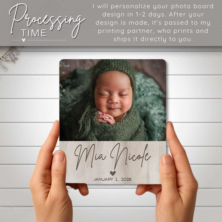 Person holding a photo board with a baby's name and date printed on it, against a light wooden background.