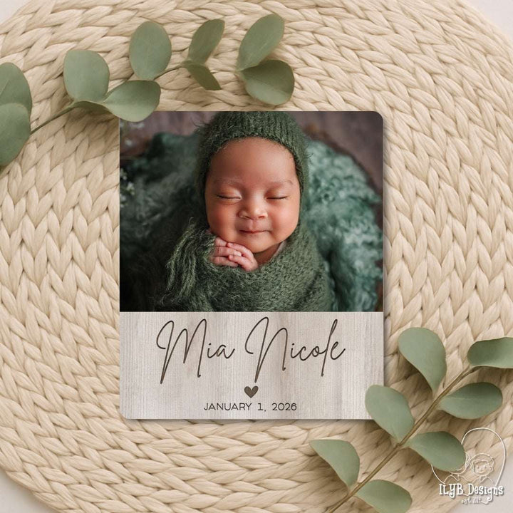 Newborn baby in green wrap and hat with 'Mia Nicole' and date on wooden plaque, surrounded by green leaves on woven surface.
