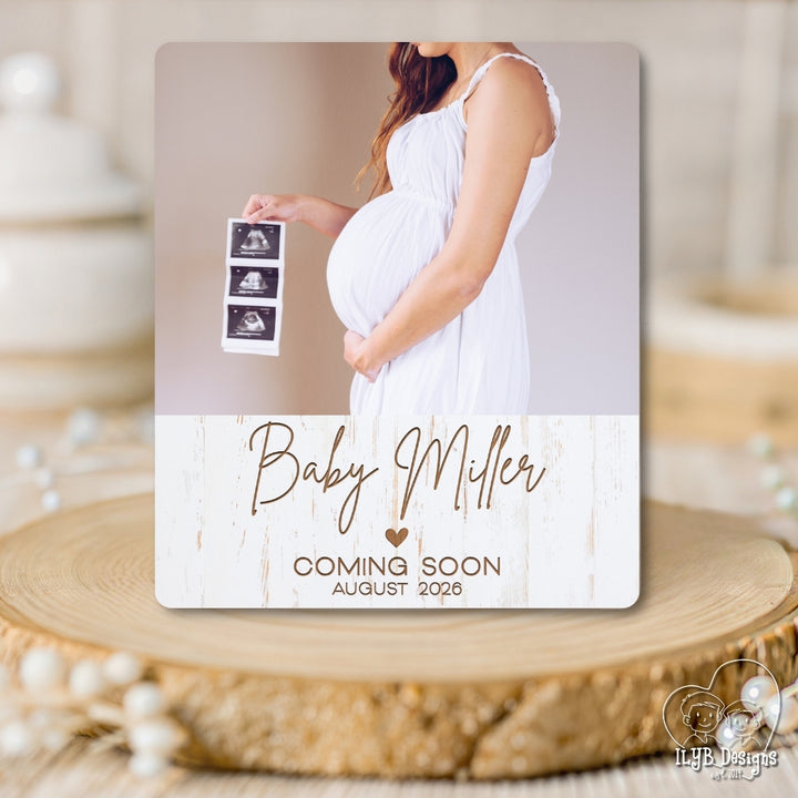 Pregnant woman holding ultrasound images with 'Baby Miller Coming Soon August 2023' text on a wooden surface.