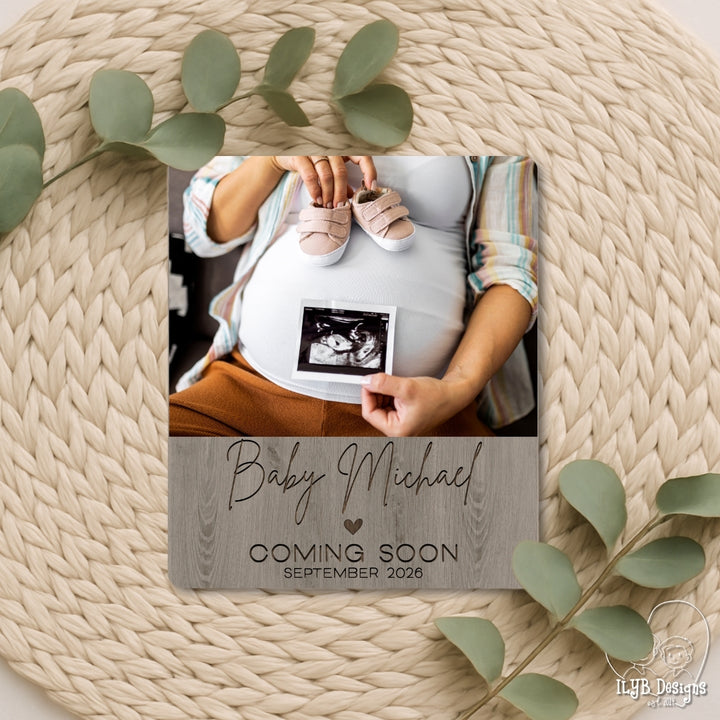 Pregnant woman holding baby shoes and ultrasound photo with 'Baby Michael' photo board announcement on a woven surface.