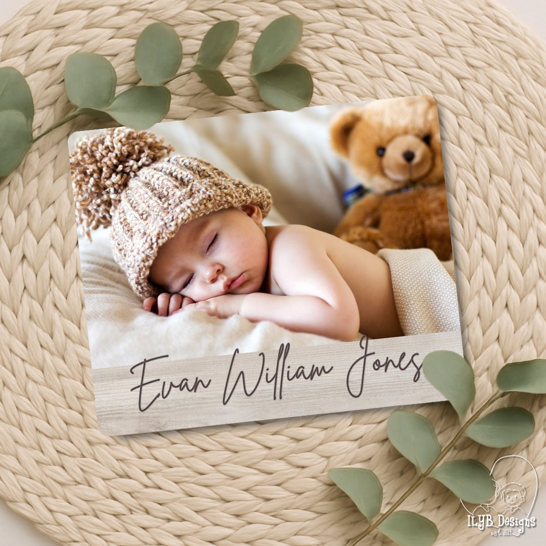 Newborn baby in a knitted hat with a teddy bear, lying on a wooden board with engraved name, surrounded by green leaves.