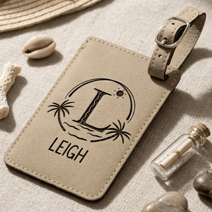 Light brown leather luggage tag with 'LEIGH' and a beach logo engraved in black, laying on a textured surface with seashells and a bottle.