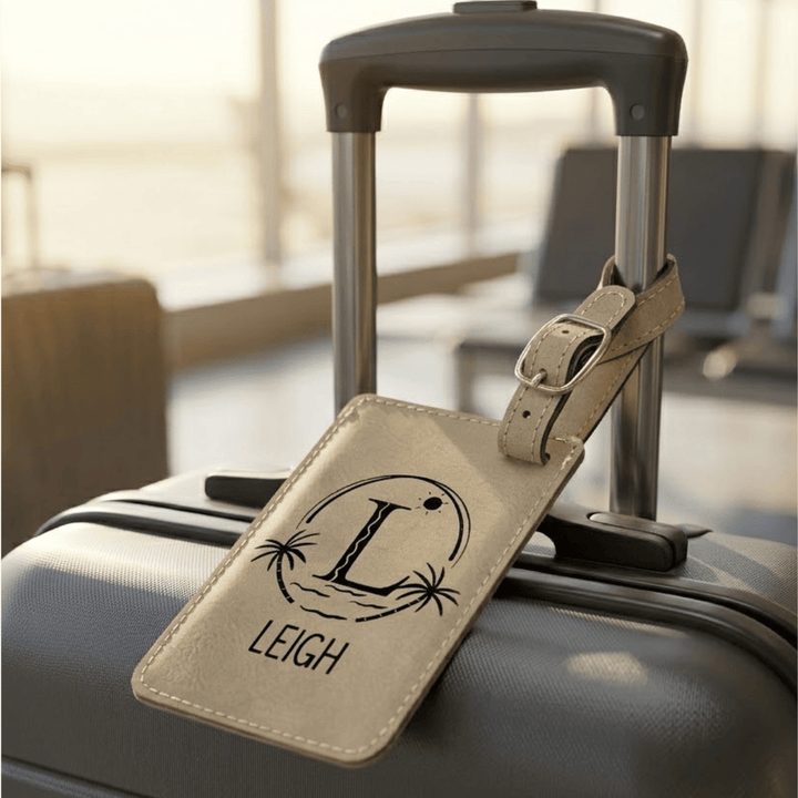 Light brown luggage tag with a beach monogram and name engraved in black, attached to a suitcase in an airport setting
