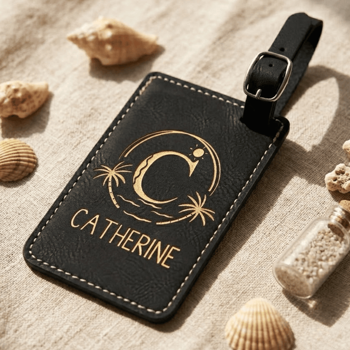 Black leather luggage tag with gold engraving and 'Catherine' text, surrounded by seashells on a textured surface.