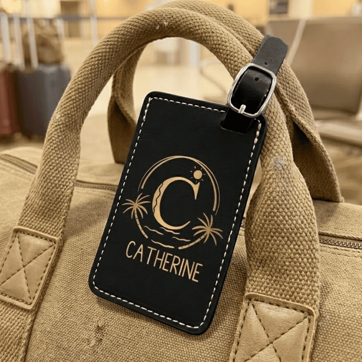 Black luggage tag with 'Catherine' and a beach logo engraved in black and attached to a beige bag.