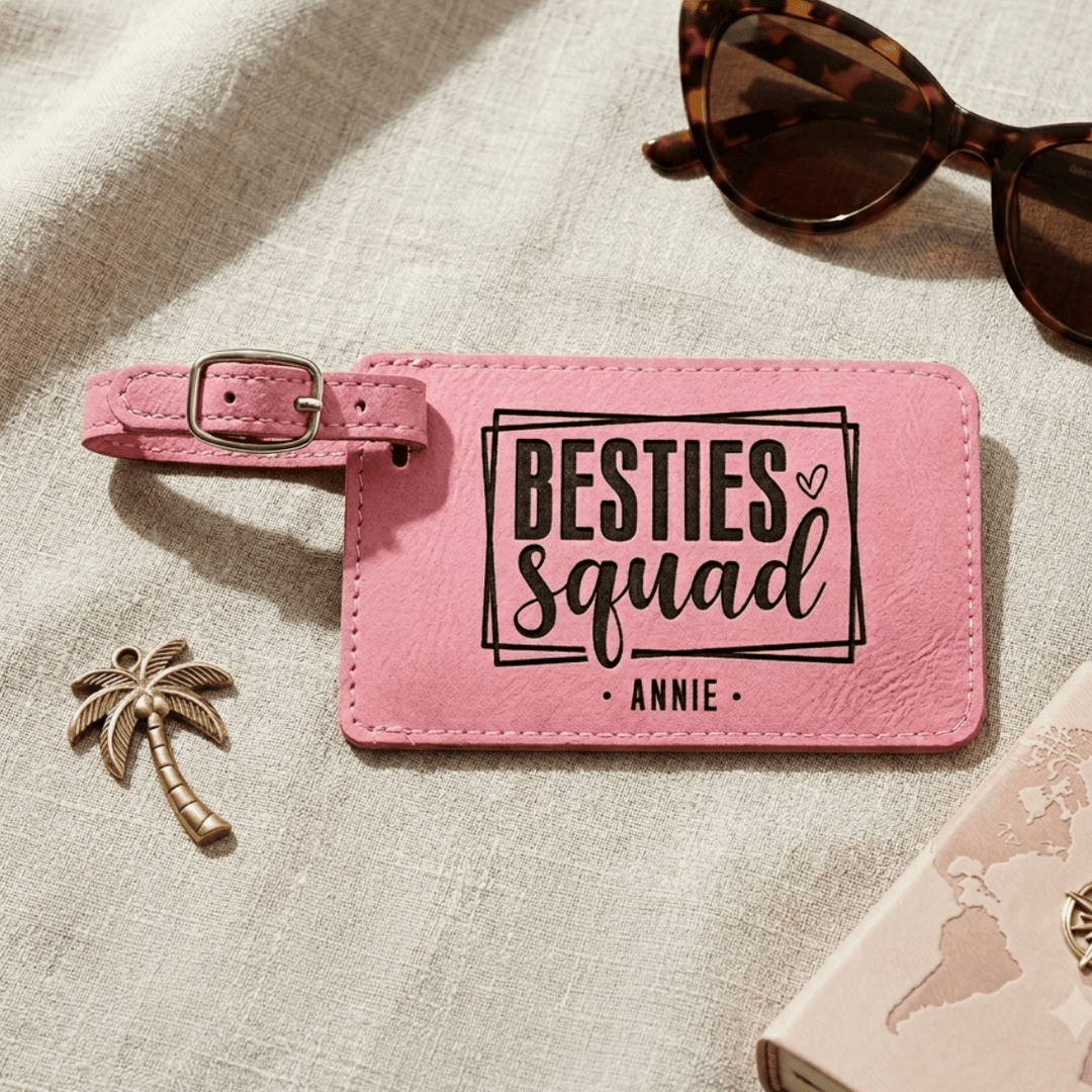 Pink luggage tag with 'BESTIES SQUAD ANNIE' text on a textured surface with sunglasses and a palm tree charm.
