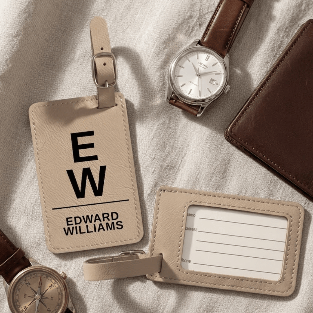 Light brown leather luggage tags with 'EW' and 'Edward Williams' engraved text, watch, and wallet on a textured surface.