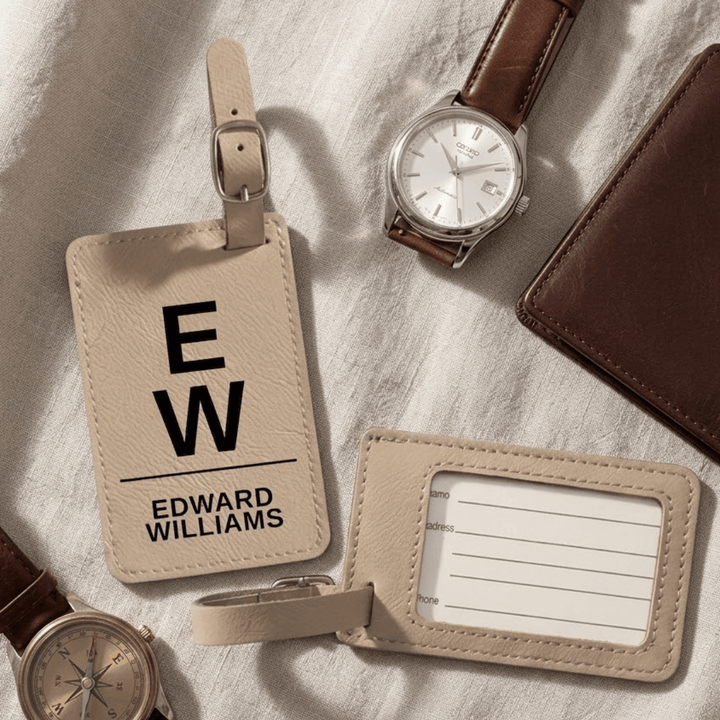 Light brown leather luggage tags with 'EW' and 'Edward Williams' engraved text, watch, and wallet on a textured surface.
