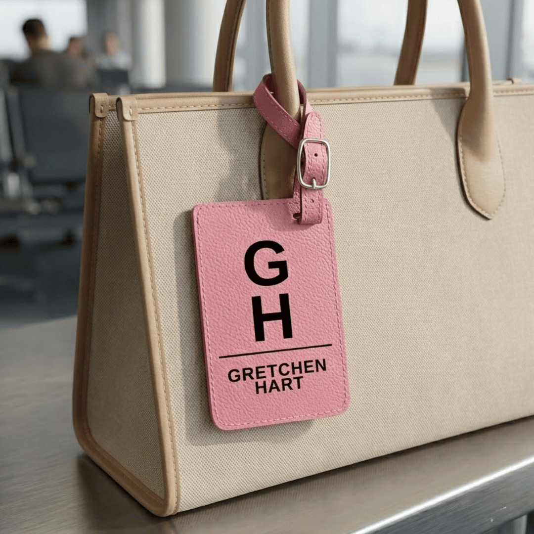 Beige handbag with a pink luggage tag featuring 'Gretchen Hart' monogram engraved in black, sitting in an airport setting.