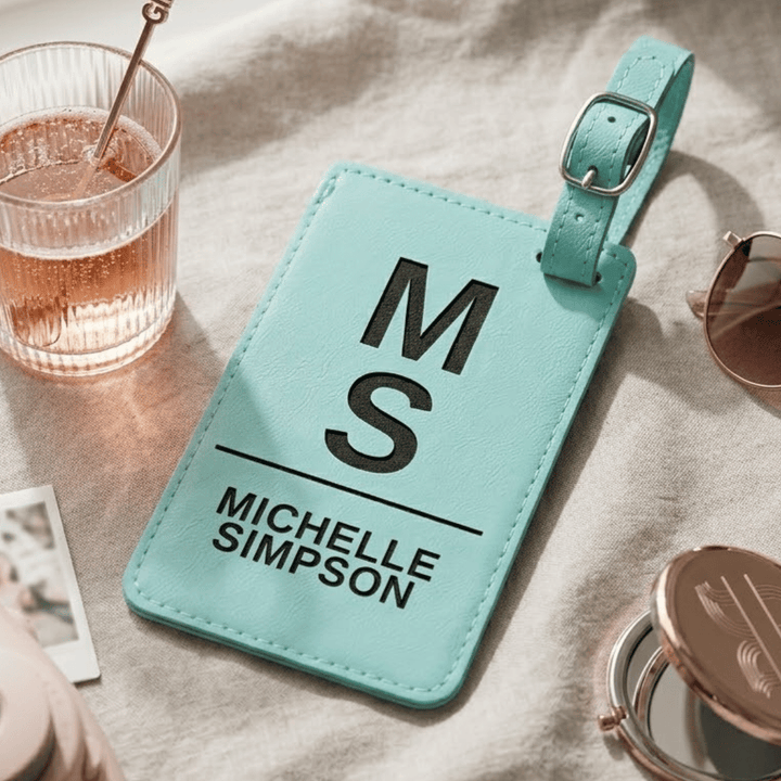 Teal luggage tag with 'MS' and 'Michelle Simpson' black engraved text on a table with a glass of water and sunglasses.