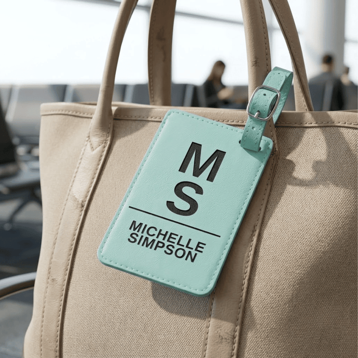 Beige bag with a turquoise luggage tag displaying 'M S' and 'Michelle Simpson' engraved in black in an airport setting.
