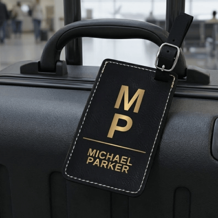 Black luggage tag with gold lettering engraved monogram attached to a suitcase in an airport setting