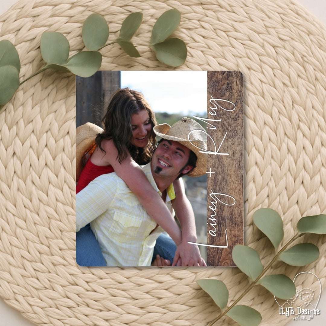 Wooden photo frame with a couple's photo and 'Lasley & Kevy' text, surrounded by green leaves on a woven surface.