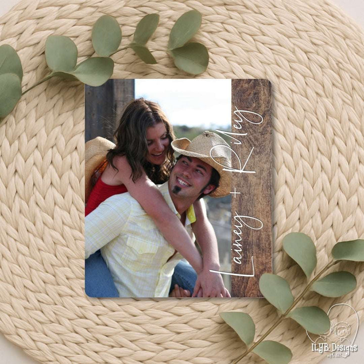 Wooden photo frame with a couple's photo and 'Lasley & Kevy' text, surrounded by green leaves on a woven surface.