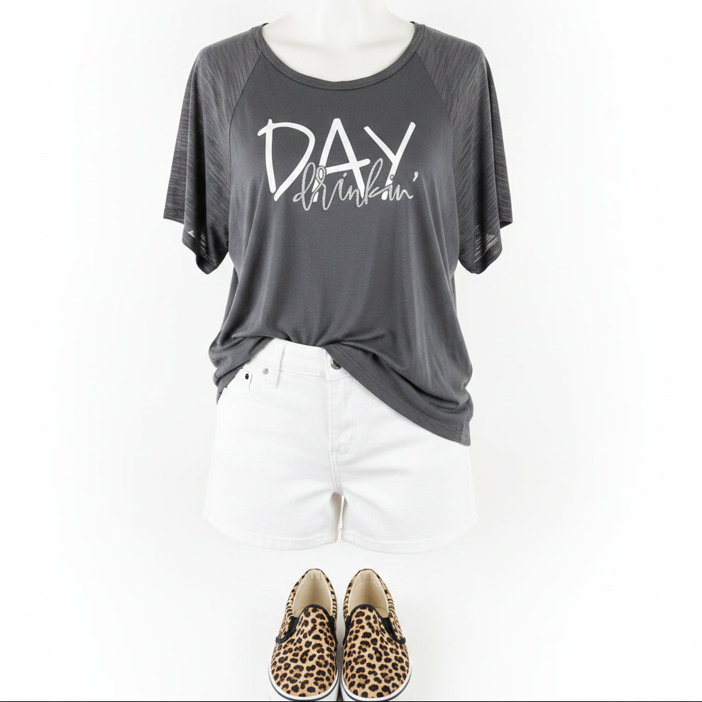 A dark gray raglan t-shirt with 'DAY drinkin' printed in white and silver glitter on the front, styled on a mannequin with white shorts and leopard print slip-on shoes against a white background.