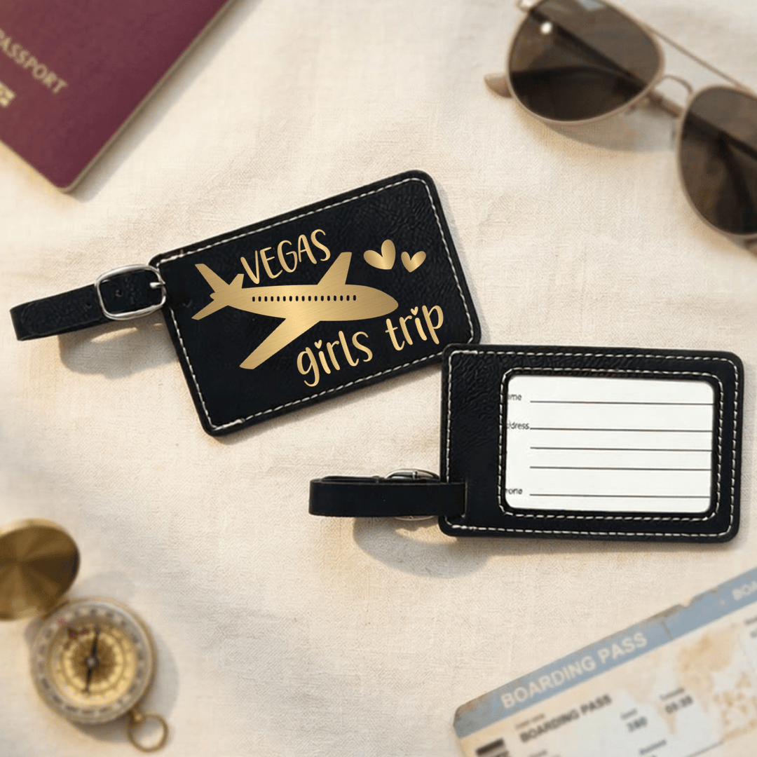 Black luggage tag with 'Vegas girls trip' text and airplane design on a textured surface with travel items.