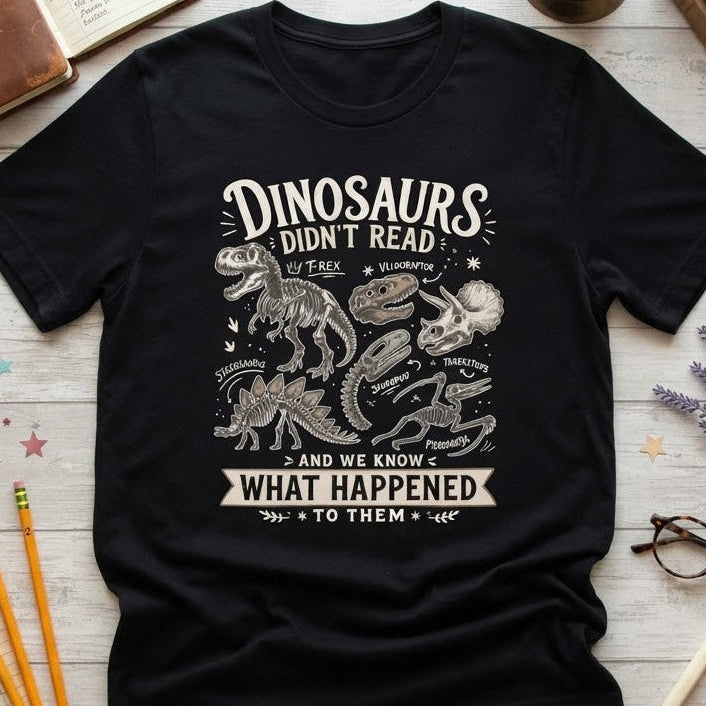 A black graphic t-shirt laid flat on a light wood background, surrounded by school and study props including books, pencils, glasses, and a brass object. The shirt features a white distressed graphic that reads 'DINOSAURS DIDN'T READ' with dinosaur illustrations and the phrase 'AND WE KNOW WHAT HAPPENED TO THEM'.