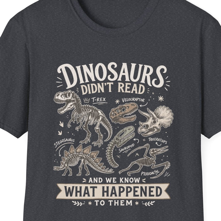 A dark charcoal gray short-sleeve t-shirt with a vintage-style dinosaur graphic on the front. The graphic features white and light beige text reading 'DINOSAURS DIDN'T READ' and 'AND WE KNOW WHAT HAPPENED TO THEM', surrounded by detailed illustrations of dinosaur skeletons and a T-Rex.