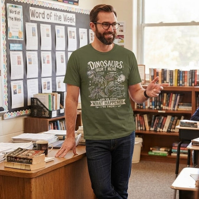 A man wearing a green graphic t-shirt that reads 'DINOSAURS DIDN'T READ AND WE KNOW WHAT HAPPENED TO THEM' stands in a classroom, gesturing with his hand. The room has bookshelves, a whiteboard, and other students in the foreground.