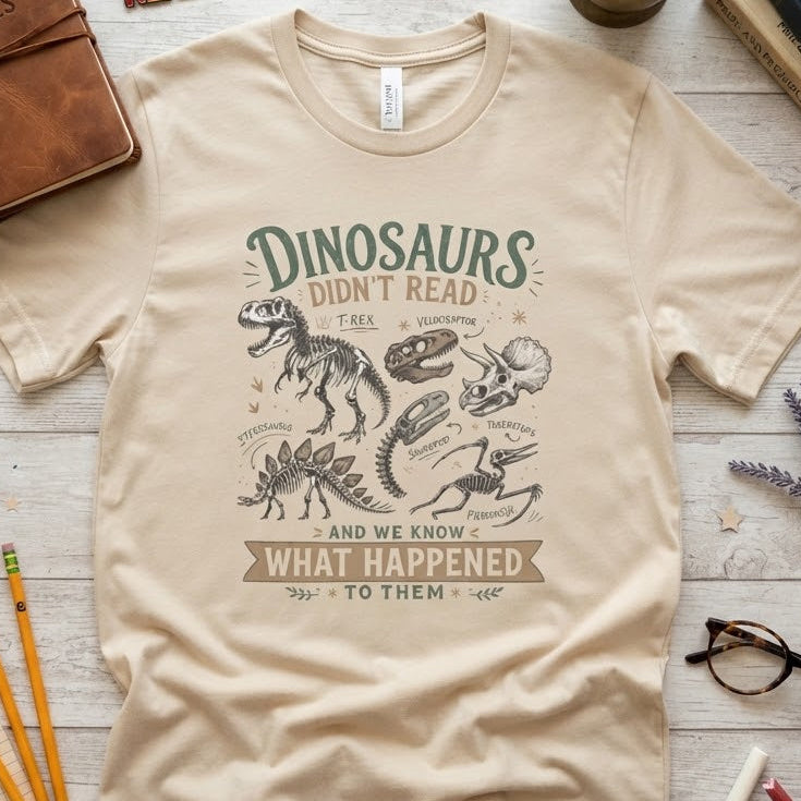 A beige graphic t-shirt featuring dinosaur illustrations and the text 'Dinosaurs Didn't Read and we know What Happened To Them' in green and brown lettering. The t-shirt is displayed in a flat-lay arrangement on a light wood background, surrounded by educational props including a leather notebook, books, pencils, glasses, and lavender.
