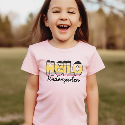 Young girl wearing light pink personalized school shirt that says hello in pencil shaped letters and kindergarten in black script underneath.