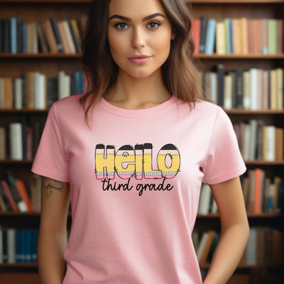 Adult woman wearing light pink personalized teacher shirt with hello in pencil letters and third grade in black script underneath.