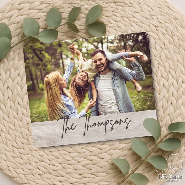 Family photo on a wooden plaque with 'The Thompsons' on a woven surface with green leaves.