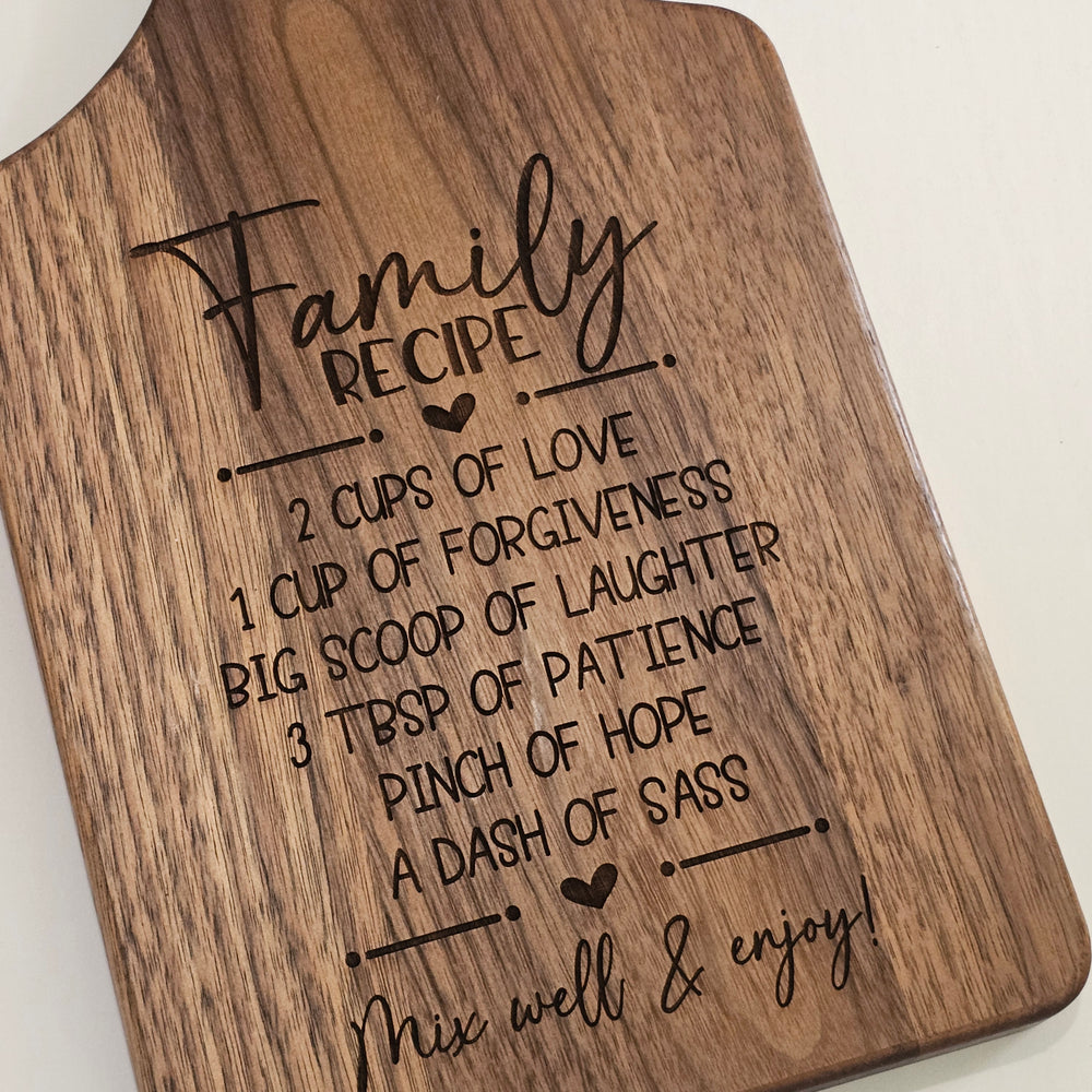 A close-up of a wooden cutting board with rounded corners, featuring a laser-engraved 'Family Recipe' print with humorous cooking instructions and the phrase 'Mix, well & enjoy!' on a light neutral background.