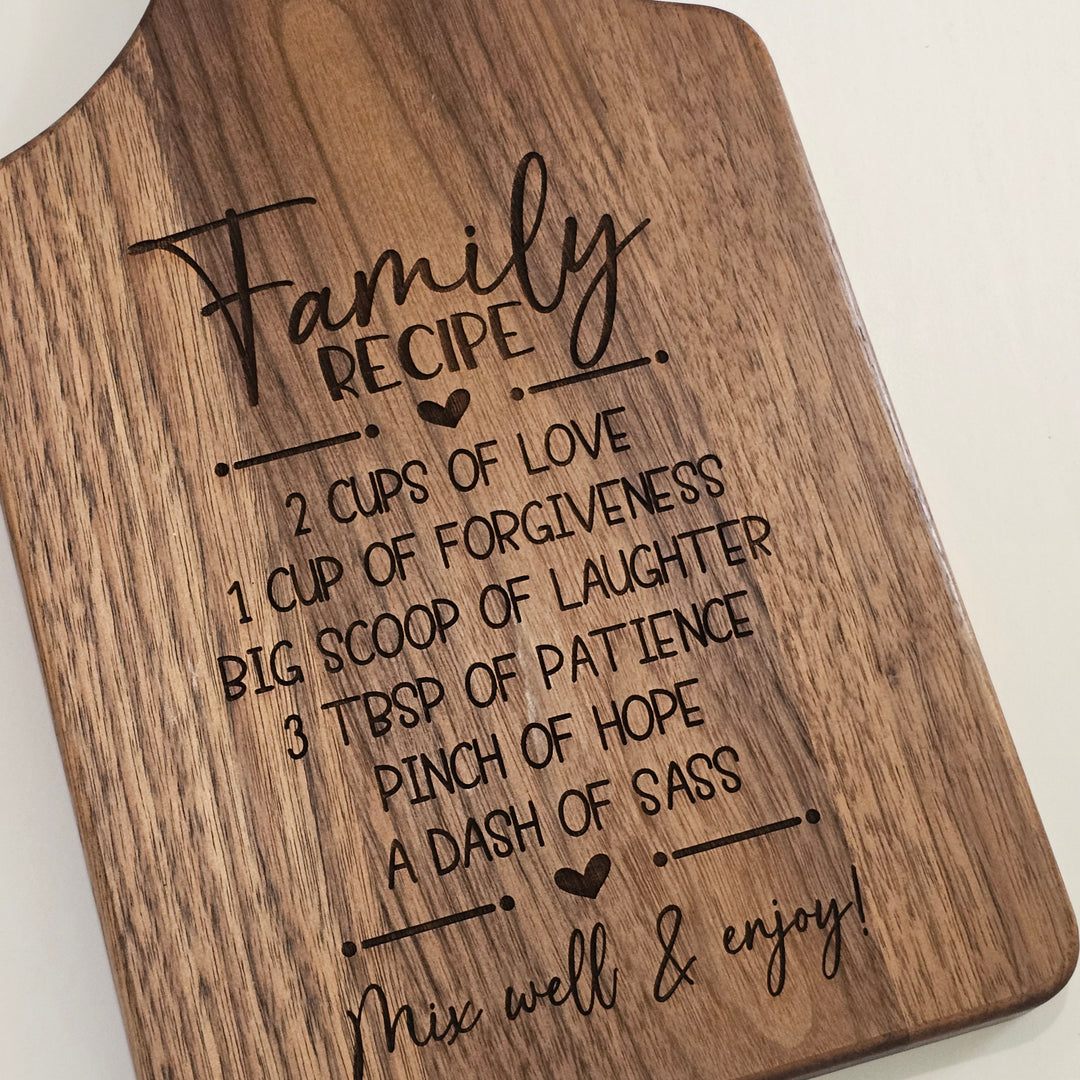 A close-up of a wooden cutting board with rounded corners, featuring a laser-engraved &