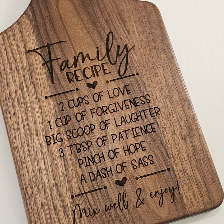 A close-up of a wooden cutting board with rounded corners, featuring a laser-engraved &