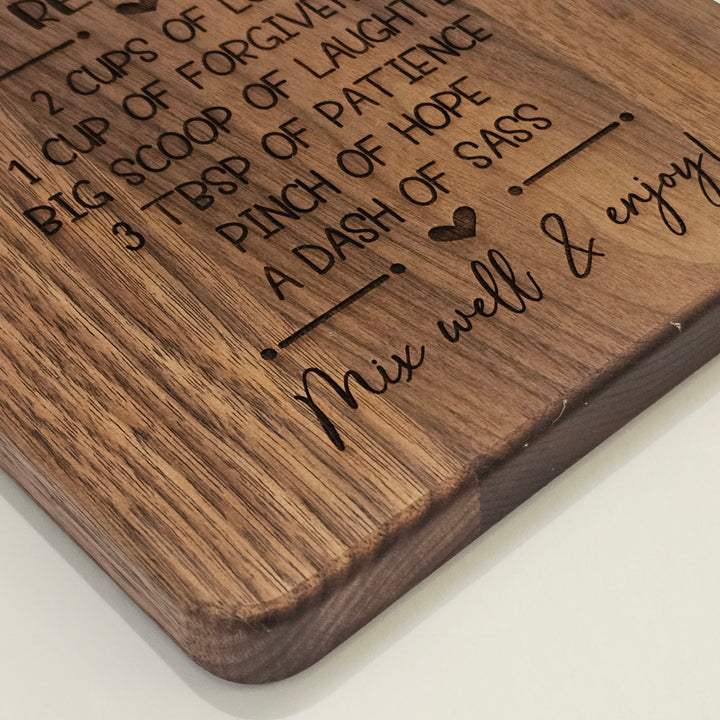 A close-up of a rectangular wooden cutting board with rounded corners, featuring engraved text and a humorous recipe-style design on a light neutral background.