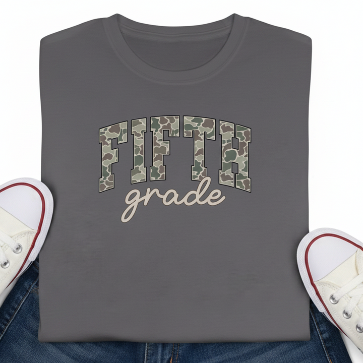 A folded dark charcoal gray t-shirt with 'FIFTH grade' printed on the front. The word 'FIFTH' is filled with a green, brown, and beige camouflage pattern, while 'grade' is in a light beige script.