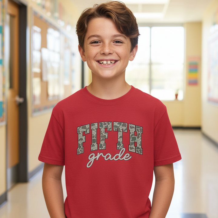 A young boy smiling in a school hallway, wearing a red t-shirt with 'FIFTH grade' printed on the front in camouflage and tan script.