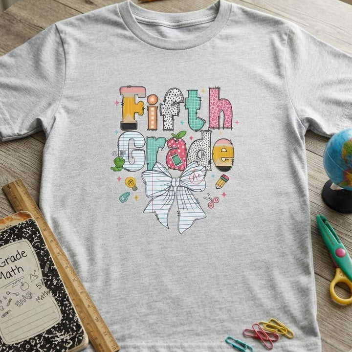 A light ash gray heather t-shirt with a colorful 'Fifth Grade' graphic featuring a bow and school-themed doodles, styled in a flat-lay on a wooden desk with school supplies including a notebook, pencil, scissors, and a globe.