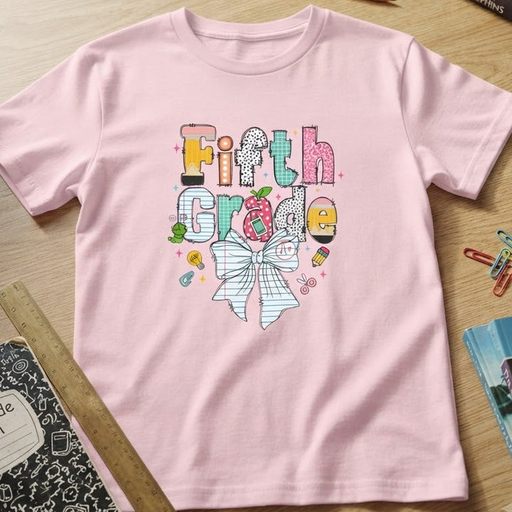 A light pink short-sleeve t-shirt with a colorful 'Fifth Grade' graphic and a striped bow design, laid flat on a wooden desk surrounded by school supplies and books.