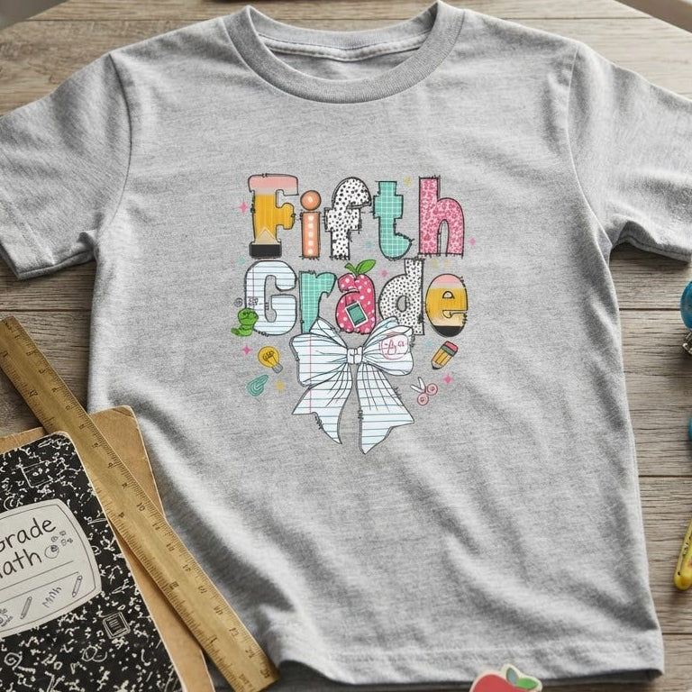 A heather gray short-sleeve t-shirt with a colorful 'Fifth Grade' graphic and a white bow design, styled in a flat-lay on a wooden table with school supplies including a notebook, pencil, ruler, and scissors.