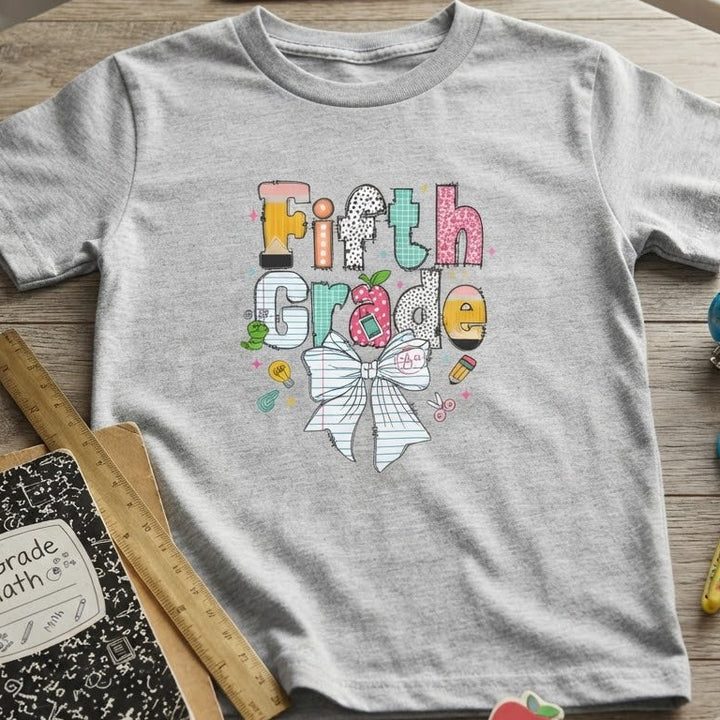 A heather gray short-sleeve t-shirt with a colorful 'Fifth Grade' graphic and a white bow design, styled in a flat-lay on a wooden table with school supplies including a notebook, pencil, ruler, and scissors.