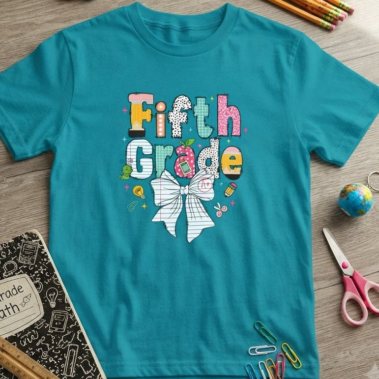 A turquoise short-sleeve t-shirt with a colorful 'Fifth Grade' bow graphic, styled in a flat lay on a gray wood-grain surface with school supplies including pencils, a notebook, scissors, and paper clips.